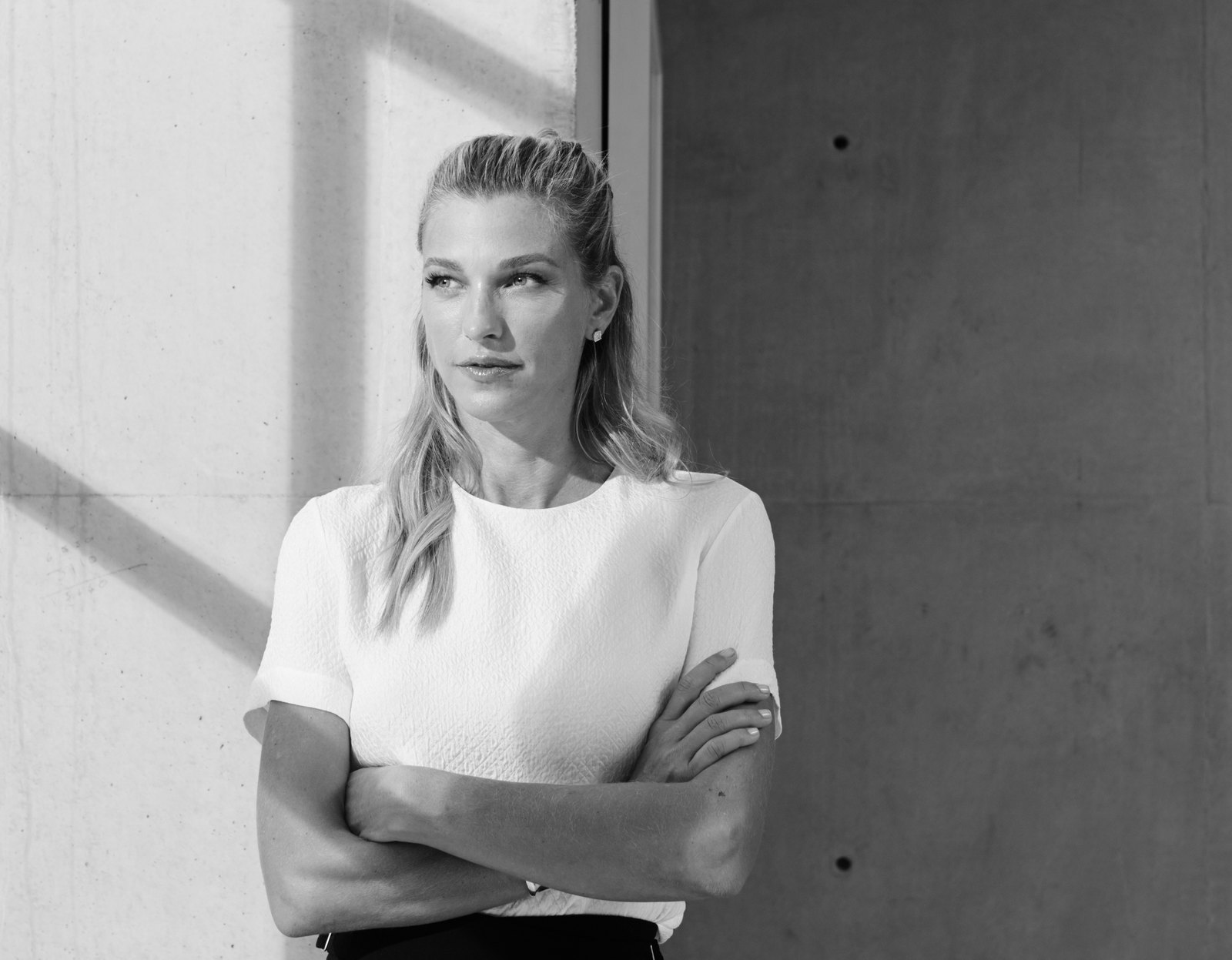 Black and white editorial portrait of Nicole Junkerman standing by a sunlit concrete wall.
